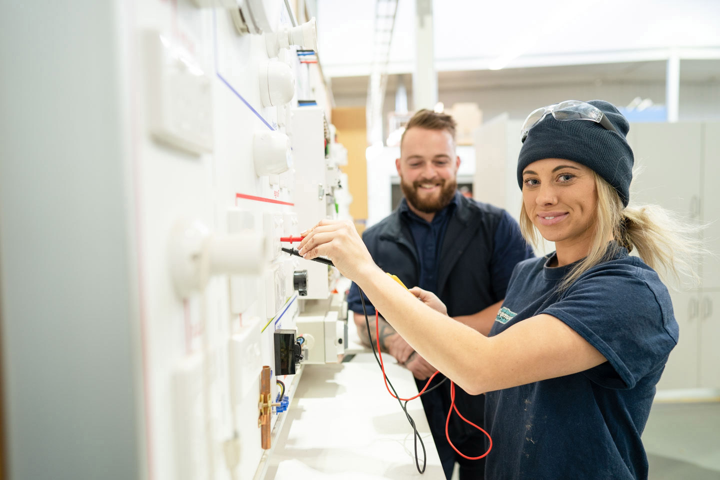 Young female Electrotechnology Apprentice participating in hands-on learning at an electrical switchboard training aid, whilst being overseen by her educator at TAFE