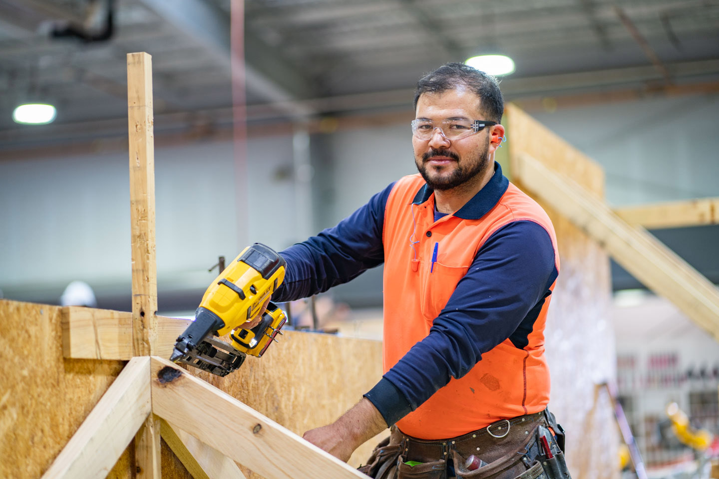 Carpentry Apprentice participating in hands-on learning at TAFE. He is dressed in high-vis and appropriate PPE, and is holding a nail-gun.