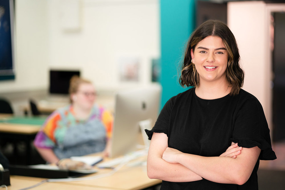 Young Female student smiling at the camera. Design student blurred in the background learning on her Apple Mac.