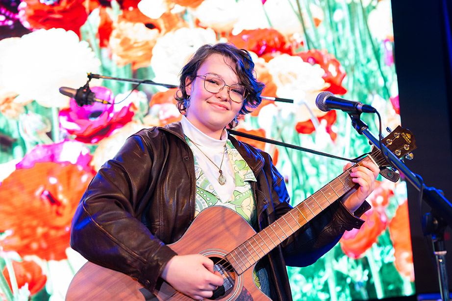 Young Female student studying Music Industry and Sound Production, playing the guitar with a microphone in front of her and a vibrantly coloured backdrop